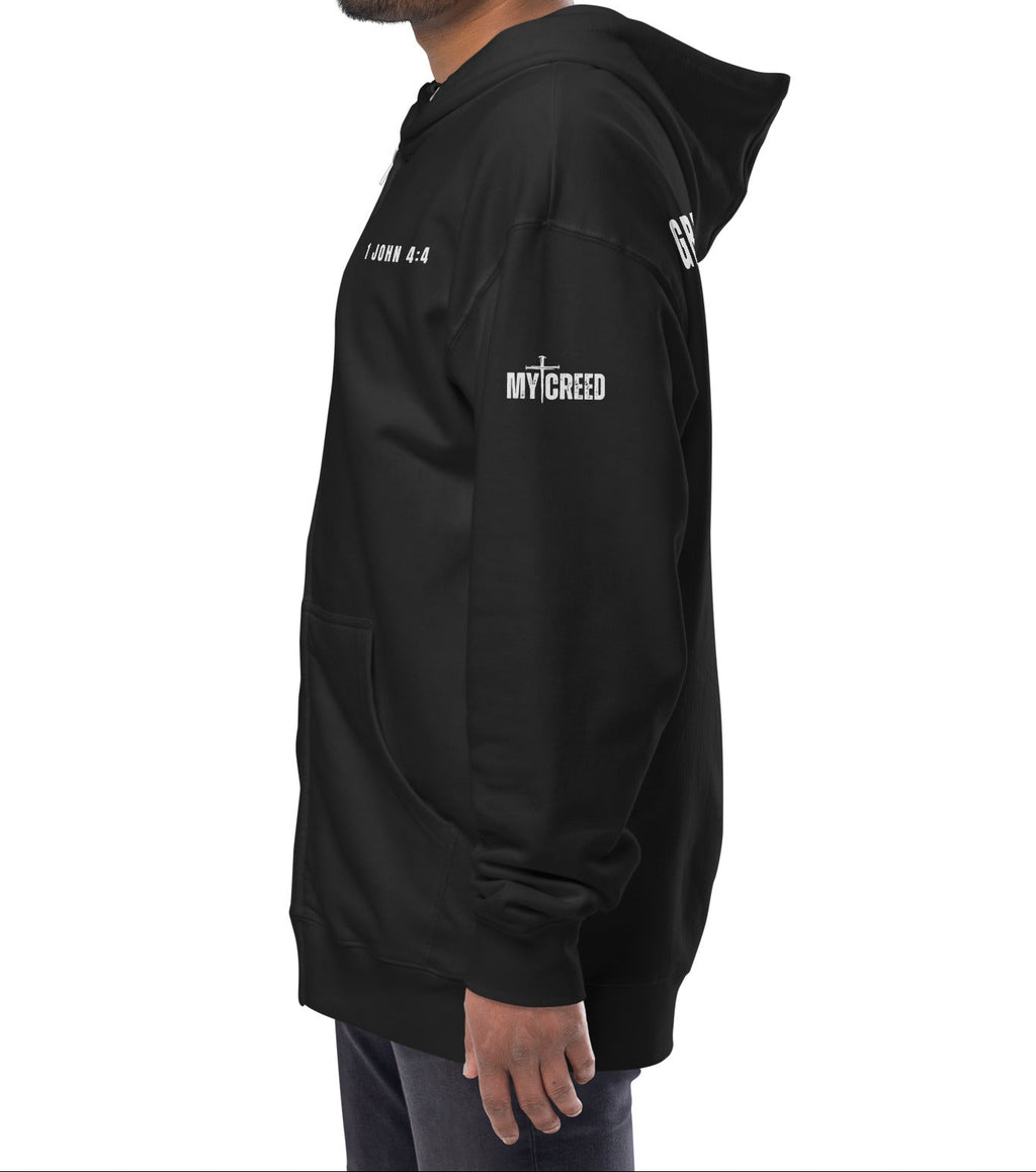 Black zip-up hoodie on model, angled back/side view of “GREATER IS HE — 1 JOHN 4:4” by MyCreed™
