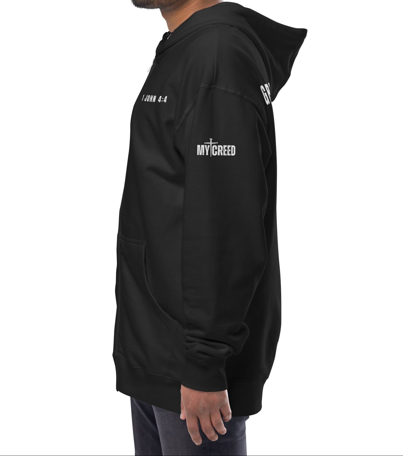 Black zip-up hoodie on model, angled back/side view of “GREATER IS HE — 1 JOHN 4:4” by MyCreed™